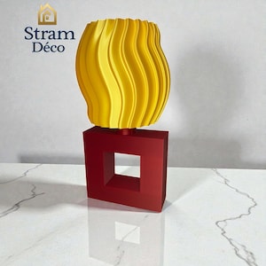 Puede incluir: Una escultura decorativa moderna con una parte superior ondulada de color amarillo vibrante y una base cuadrada roja audaz con un recorte central. La escultura se exhibe sobre una superficie blanca. El texto "Stram Déco" es visible.