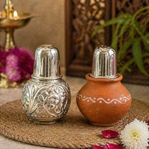 May include: Two decorative salt and pepper shakers. One is silver with an embossed floral design, the other is terracotta with a white dotted pattern. Both have silver lids. They sit on a woven mat with rose petals.