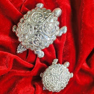 Puede incluir: Dos figuras de tortuga en tono plateado con intrincados patrones de volutas en relieve en sus caparazones. La tortuga más grande está colocada sobre la más pequeña, ambas con cabezas, patas y colas detalladas con un acabado metálico.