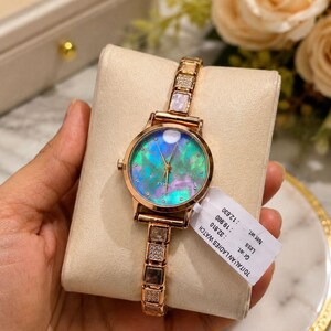 May include: A rose gold ladies' watch with an iridescent face and a matching band. The watch face has a pearlescent effect with blue, green, and pink hues. The band features square, gem-studded links. The watch is displayed on a cream-coloured cushion.