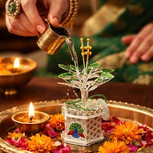May include: A silver and glass decorative tree is being watered from a small brass pitcher. The tree sits in a silver box with a peacock design, surrounded by orange flowers and candles on a brass tray. A hand with a ring and bracelets is visible.