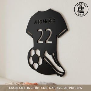 Könnte beinhalten: Schwarze, lasergeschnittene Wandkunst im Fußball-Design. Das Design zeigt ein Trikot mit dem Namen "ALEXANDER" und der Nummer "22". Ein Fußball und ein Fußballschuh sind ebenfalls enthalten. Der Text "LASER CUT FILES" befindet sich in einem Kreis. Der untere Text lautet "LASER CUTTING File : CDR, DXF, SVG, AI, PDF, EPS".