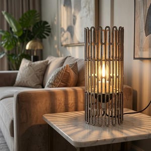 May include: A cylindrical table lamp featuring a dark brown wooden frame with a repeating vertical cutout pattern. The lamp houses a single glowing light bulb inside, casting linear shadows across the surface. A black power cord extends to a wall outlet.
