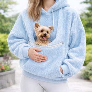 Op de afbeelding: Een lichtblauwe, pluizige hoodie met een kangoeroezak om een kleine hond in te dragen. Een bruin-beige Yorkshire Terrier is zichtbaar in de zak. De hoodie heeft een capuchon met trekkoord en geribde manchetten.