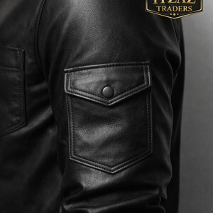 Op de afbeelding: Close-up van een zwarte leren jas met een zak met knoop op de mouw. De jas heeft een klassiek ontwerp met een kraag en een knoopsluiting. Het logo "REAL LEATHER ITZAZ TRADERS" is zichtbaar.