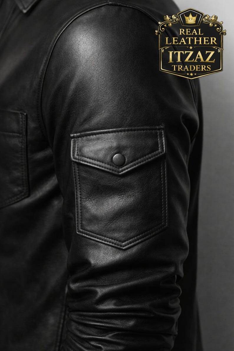 Op de afbeelding: Close-up van een zwarte leren jas met een zak met knoop op de mouw. De jas heeft een klassiek ontwerp met een kraag en een knoopsluiting. Het logo "REAL LEATHER ITZAZ TRADERS" is zichtbaar.