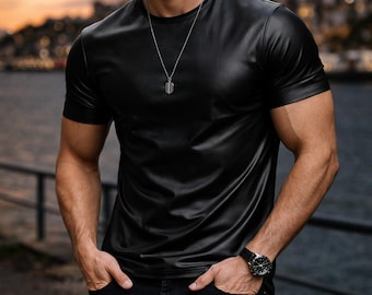 Op de afbeelding: Zwart leren T-shirt met korte mouwen. Het shirt wordt gedragen met een zwarte jeans en een zilveren ketting. De afbeelding toont ook een zwart horloge en een zonnebril. De tekst "REAL LEATHER" en "ITZAZ TRADERS" zijn zichtbaar.