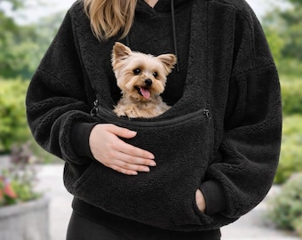 May include: Black hooded sweatshirt with a kangaroo pocket designed to carry a small dog. The dog is a light brown and white Yorkshire Terrier. The sweatshirt is made of a soft, textured material and is worn with black shorts.