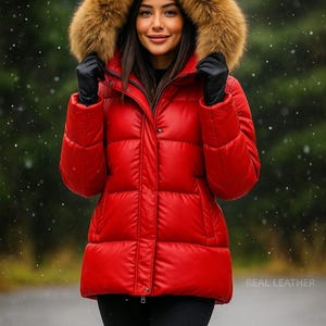 May include: A red puffer jacket with a fur-trimmed hood. The jacket has a quilted design and a zip closure. The model is wearing black gloves and black trousers. The text "REAL LEATHER" is visible on the lower right.