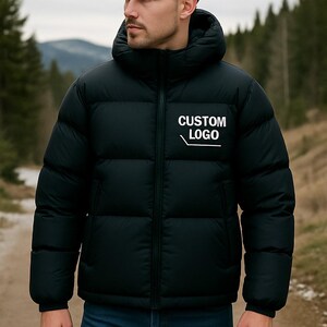 Può includere: Giacca puffer nera con cappuccio e chiusura a zip. La giacca presenta il testo bianco "CUSTOM LOGO" stampato sul petto. La persona indossa jeans blu. Lo sfondo è una scena all'aperto sfocata.