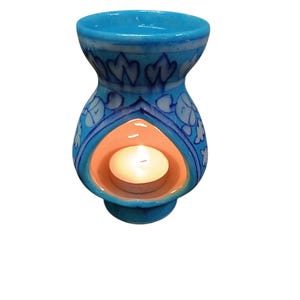 May include: A teal ceramic essential oil diffuser with a floral design. The diffuser has a round base, a central opening for a tea light, and a top bowl. A lit tea light is inside, creating a warm glow. Home decor.