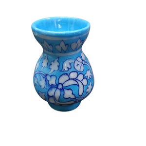 Könnte beinhalten: Eine kleine, türkisfarbene Keramikvase mit rundem Körper und ausgestelltem Rand. Die Vase ist mit einem weißen Blumenmuster verziert, das mit Dunkelblau umrandet ist. Das Design zeigt stilisierte Blumen und Blätter und erzeugt einen dekorativen und künstlerischen Look.