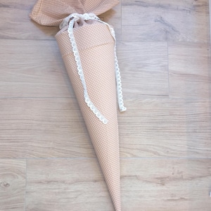 May include: A cone-shaped fabric gift bag in a light tan color with a small white polka dot pattern. The top of the bag is gathered and tied with a white lace ribbon that hangs down the sides, creating a decorative closure for the textile container.