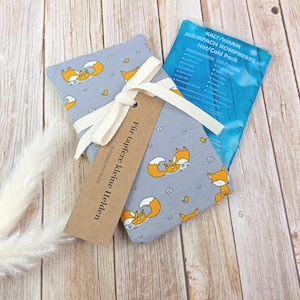 Könnte beinhalten: Eine graue Stoffhülle mit einem Muster aus orangefarbenen Füchsen und kleinen Blumen, gebunden mit einem cremefarbenen Band. Ein braunes Etikett trägt die Aufschrift "Für tapfere kleine Helden" und ein blaues Wärme-/Kältepack ist sichtbar.