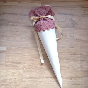 May include: A cone-shaped gift bag featuring a solid white lower section and a dusty rose upper section with a small floral pattern. The top is gathered and secured with a light beige satin ribbon tied in a bow, creating a decorative closure.