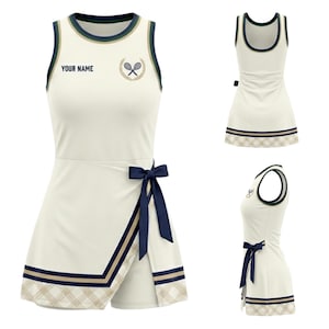 May include: Cream tennis dress with navy and gold trim. It has a wrap skirt with a navy bow, and the text "YOUR NAME" above a tennis racket and laurel wreath emblem. The dress has a sleeveless design.