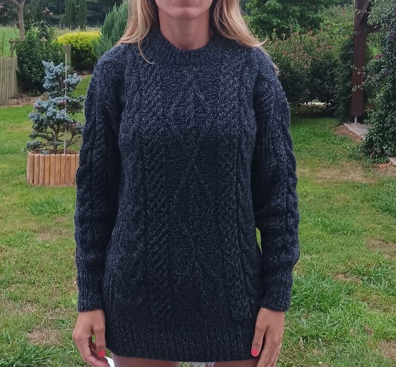 May include: A dark gray, cable-knit sweater with a crew neck. The sweater has a textured pattern and long sleeves. The person is standing in a grassy outdoor setting.