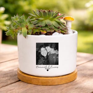 May include: A white ceramic planter with a succulent plant and a small mushroom figurine. The planter features a black and white photo with the names "Hannah & Lucas" and the date "06.11.2025". The planter sits on a round wooden base.