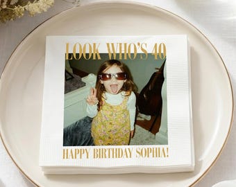 May include: A square white paper napkin featuring a printed photograph of a child wearing sunglasses and a yellow floral dress. The text "LOOK WHO'S 40" appears in gold above the photo, and "HAPPY BIRTHDAY SOPHIA!" is printed in gold below.