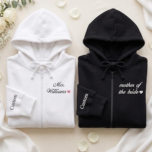 May include: A white zip-up hoodie with the text Mrs. Williams and a pink heart, and a black zip-up hoodie with the text mother of the bride and a white heart. Both hoodies feature the word Custom printed on the sleeve cuff.