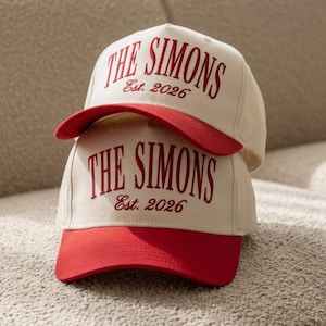 May include: Two stacked baseball caps featuring an off-white crown and a red brim. The front of each cap displays the red embroidered text THE SIMONS in a bold serif font, with the smaller script text Est. 2026 positioned directly underneath.