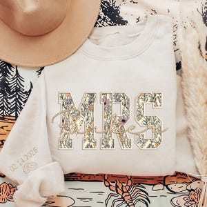 May include: Cream-colored sweatshirt with floral "MRS Parker" design. The letters are filled with a floral pattern in shades of purple, green, and yellow. The sleeve has the date "02.14.2026" embroidered on it.