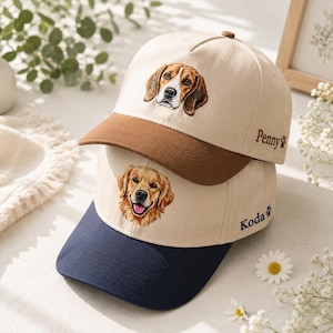 May include: Two baseball caps stacked on each other. The top cap is cream with a brown brim and an embroidered beagle, featuring the name Penny and a paw print. The bottom cap is cream with a navy brim and an embroidered golden retriever, featuring the name Koda and a paw print.
