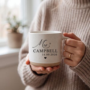 May include: A speckled off-white ceramic mug with a handle, featuring black printed text that reads Mrs CAMPBELL 14.09.2026, accented with a small heart icon above the name and a solid black heart icon centered below the date.