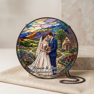 May include: A circular stained glass suncatcher featuring a bride and groom standing in a garden with a cottage and mountains in the background. The text "Brown & Amelia September 20, 2026" is printed at the bottom. It includes a black metal chain.