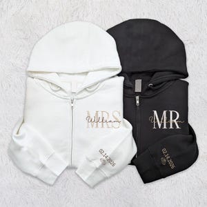May include: Two zip-up hoodies, one white and one black, are shown. The white hoodie has "MRS William" embroidered in gold script, with "02.14.2026" on the sleeve. The black hoodie features "MR" in gold script and "02.14.2026" on the sleeve.