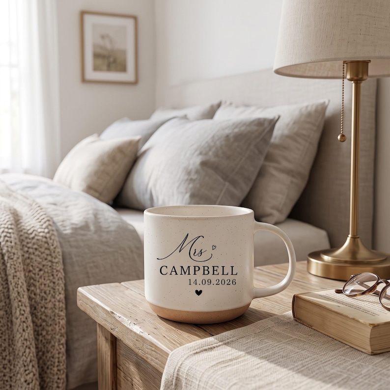 May include: A cream-colored ceramic mug with a speckled finish, featuring black printed text that reads Mrs. CAMPBELL 14.09.2026, accompanied by a small heart icon above the name and a small heart icon below the date.