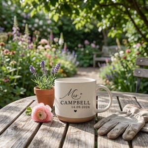 May include: A cream-colored ceramic mug with a speckled finish, featuring the black printed text Mrs. CAMPBELL 14.09.2026 and two small heart icons. The mug sits next to a small terracotta pot with lavender, a pink flower, and a pair of beige work gloves.