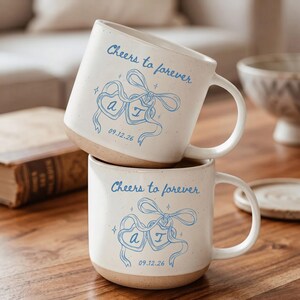 May include: Two off-white ceramic mugs with a speckled finish, each featuring blue printed text that reads Cheers to forever, two heart-shaped ornaments tied with a ribbon, and the date 09.12.26. The mugs have a smooth, rounded handle.