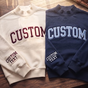 May include: Two mock neck sweatshirts, one cream with maroon lettering and one navy blue with light blue lettering. Both feature the word CUSTOM in large arched letters across the chest and the words CUSTOM TEXT embroidered on the sleeve.