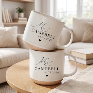 May include: Two off-white ceramic mugs with a speckled finish and a natural clay base. The top mug features black text reading Mr. CAMPBELL 14.09.2026 with a small heart. The bottom mug features black text reading Mrs. CAMPBELL 14.09.2026 with a heart.