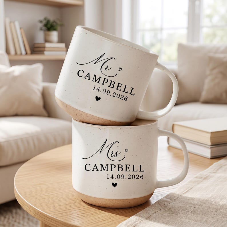 May include: Two off-white ceramic mugs with a speckled finish and a natural clay base. The top mug features black text reading Mr. CAMPBELL 14.09.2026 with a small heart. The bottom mug features black text reading Mrs. CAMPBELL 14.09.2026 with a heart.