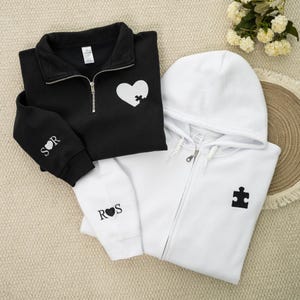 May include: Black quarter-zip sweatshirt with a white heart puzzle piece design and white hoodie with a black puzzle piece. The black sweatshirt has the initials "S R" embroidered on the sleeve. The white hoodie has the initials "R S" embroidered on the sleeve.