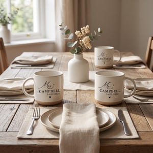 May include: Three off-white ceramic mugs with a speckled finish, each featuring black text that reads Mrs. Campbell or Mr. Campbell, the date 14.09.2026, and a small heart icon. The mugs are arranged on a wooden table with plates and napkins.