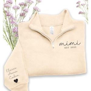 May include: Cream-colored fleece pullover with a quarter-zip neckline. The text "mimi EST 2025" is printed on the front. The left sleeve has the names "Elaina Anna Caden" and a black heart. The pullover is surrounded by purple flowers.