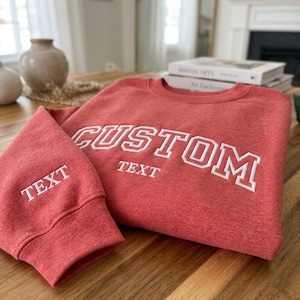 May include: A coral-colored crewneck sweatshirt featuring white embroidered lettering on the chest that reads CUSTOM TEXT, with the word TEXT also embroidered in white on the sleeve cuff. The garment has a ribbed collar, cuffs, and hem.