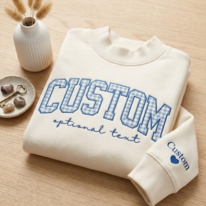 May include: A cream-colored sweatshirt featuring the word CUSTOM in blue and white gingham block letters, with the text optional text underneath. The sleeve displays the word Custom and a small blue heart icon near the ribbed cuff.