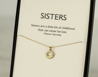 May include: A gold necklace with a circle pendant in a black gift box. The box has a white card with the text "SISTERS" and a quote about sisters. The card also includes the text "Erin Pelicano" and "14K Gold handmade USA".
