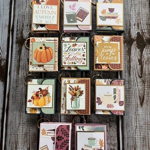 May include: A collection of miniature autumn-themed keychains. Each keychain features a small, decorative book design with fall imagery, including pumpkins, leaves, and text like "I Love Autumn" and "Leaves Are Falling". The keychains have gold-colored rings.
