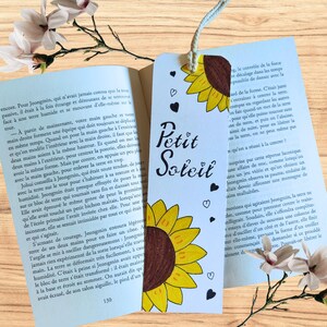 May include: A handmade bookmark with a sunflower illustration and the words "Petit Soleil" in black script. The bookmark has a white string and small heart and dot details. The background includes an open book and flowers.