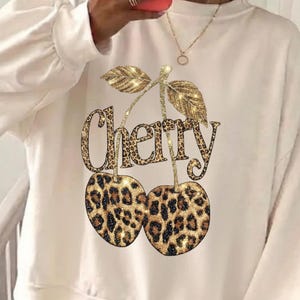 Puede incluir: Sudadera color crema con un dise&ntilde;o de cereza con estampado de leopardo brillante. La palabra "Cherry" est&aacute; escrita encima de las cerezas en un estampado de leopardo a juego. La sudadera tiene mangas largas y cuello redondo.
