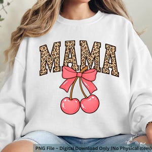 Op de afbeelding: Witte sweatshirt met het woord "MAMA" in luipaardprint letters, een roze strik en twee roze kersen. Het ontwerp is gecentreerd op de voorkant van de trui.