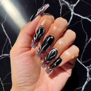 Puede incluir: Uñas acrílicas largas en forma de ataúd con un diseño degradado negro y rojo, acentuadas con detalles plateados en forma de llama. Algunas uñas presentan una pequeña gema brillante. La uña del pulgar tiene un diseño de remolino plateado.