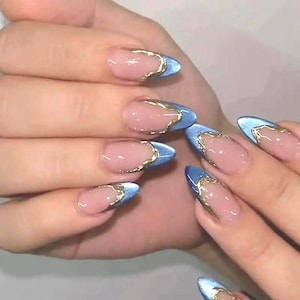 May include: Close-up of a hand with almond-shaped nails. Each nail has a light pink base, a blue tip, and a gold metallic outline. The nails are well-manicured, reflecting light, and showcase a modern nail art design.