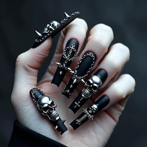 Puede incluir: Uñas postizas negras en forma de ataúd con detalles de calaveras y cruces plateadas. Las uñas tienen un acabado mate y están adornadas con cadenas y púas. Los detalles de calaveras tienen ojos rojos.