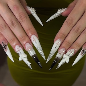 May include: Long, stiletto-shaped artificial nails with intricate designs. The nails feature white, lace, and pearl accents, with a gradient from white to black at the tips. Some nails have small cross designs and white bows.